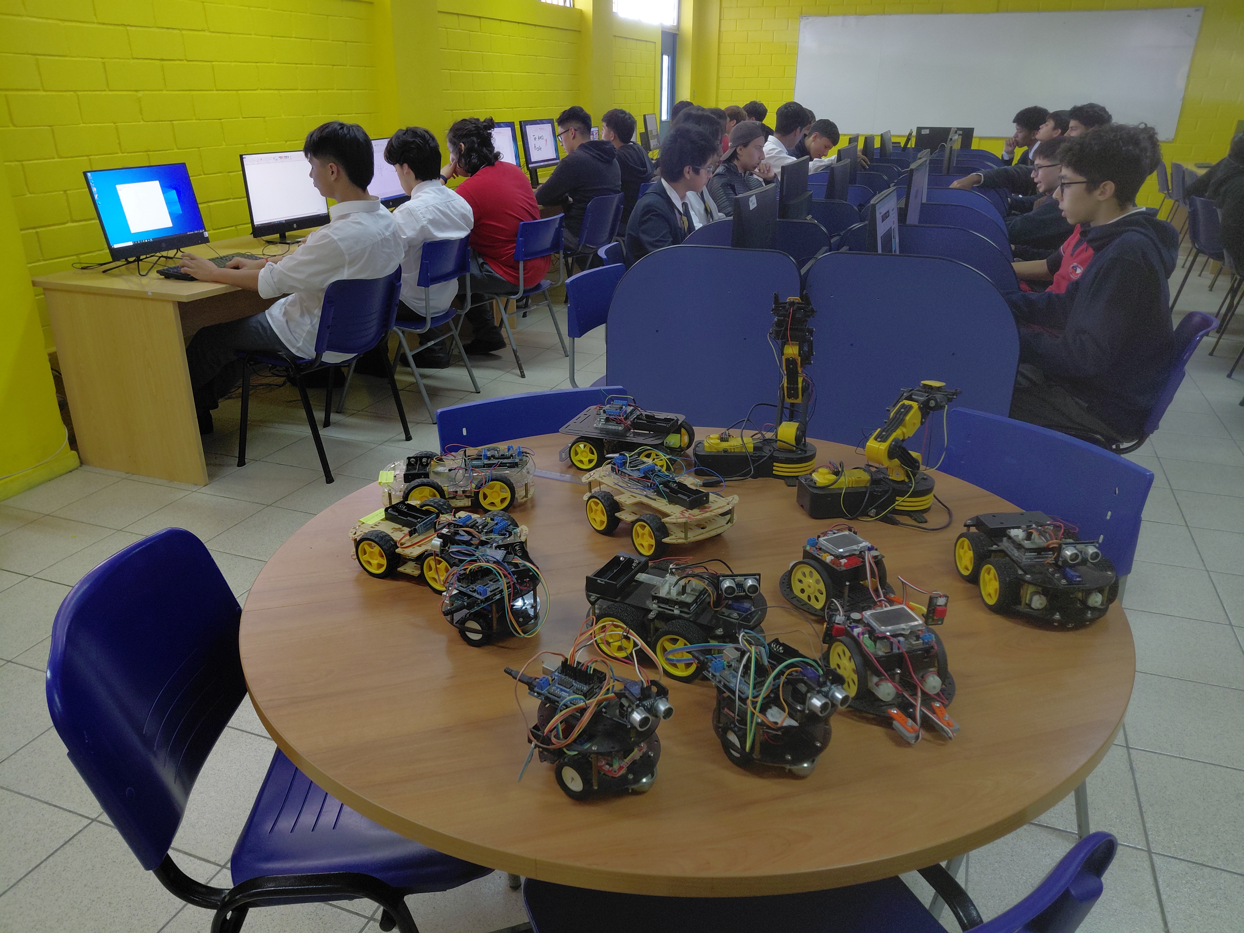 Students programming Arduino robot cars in a school robotics lab, with multiple finished robots on a central table