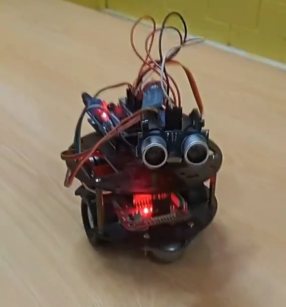 A small two-wheeled Arduino robot car with an HC-SR04 ultrasonic sensor mounted on the front, wiring visible on top