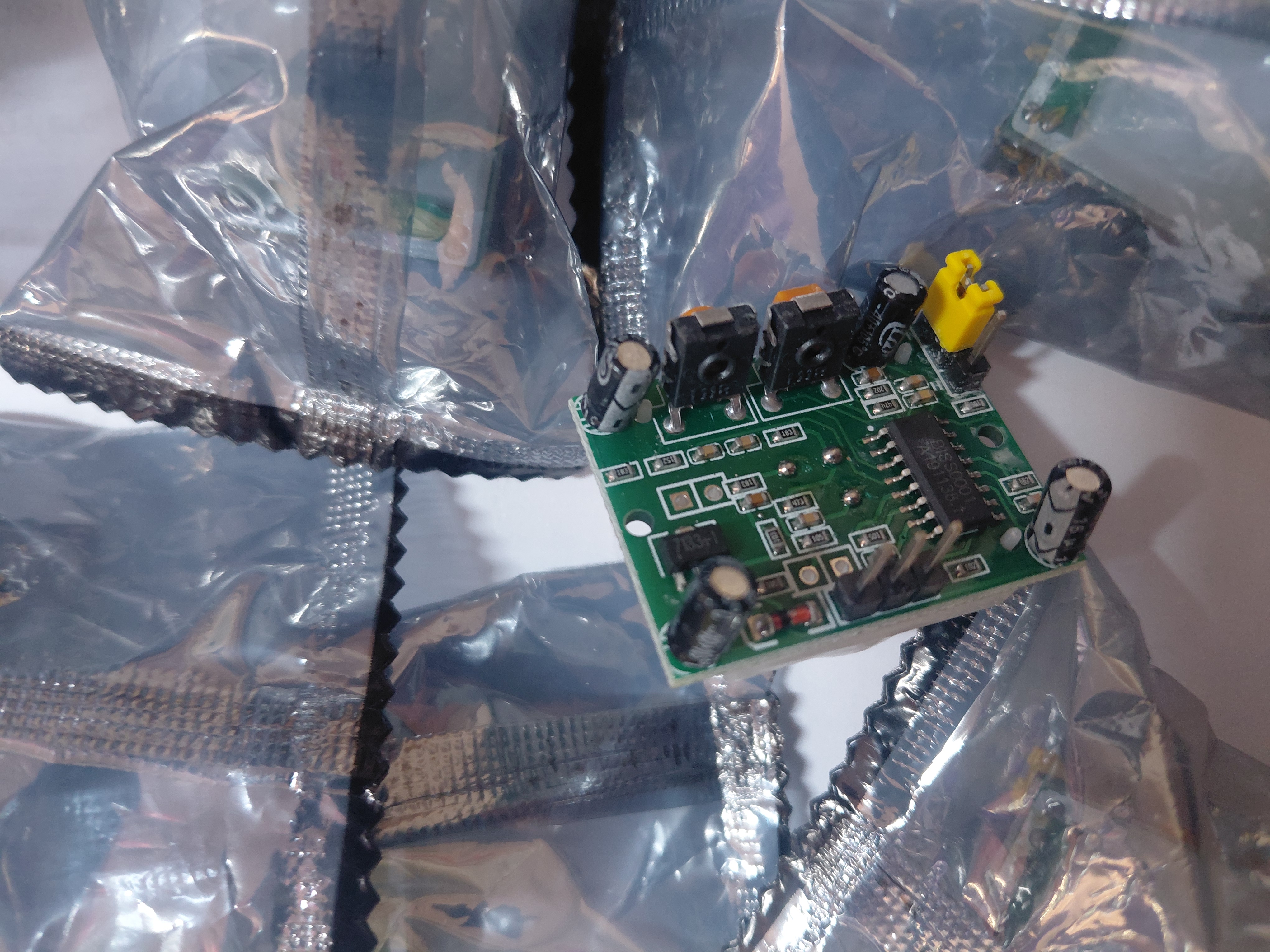 Close-up of an HC-SR501 circuit board held against antistatic bags, showing the BISS0001 controller chip, electrolytic capacitors, the three-pin header and the yellow mode jumper