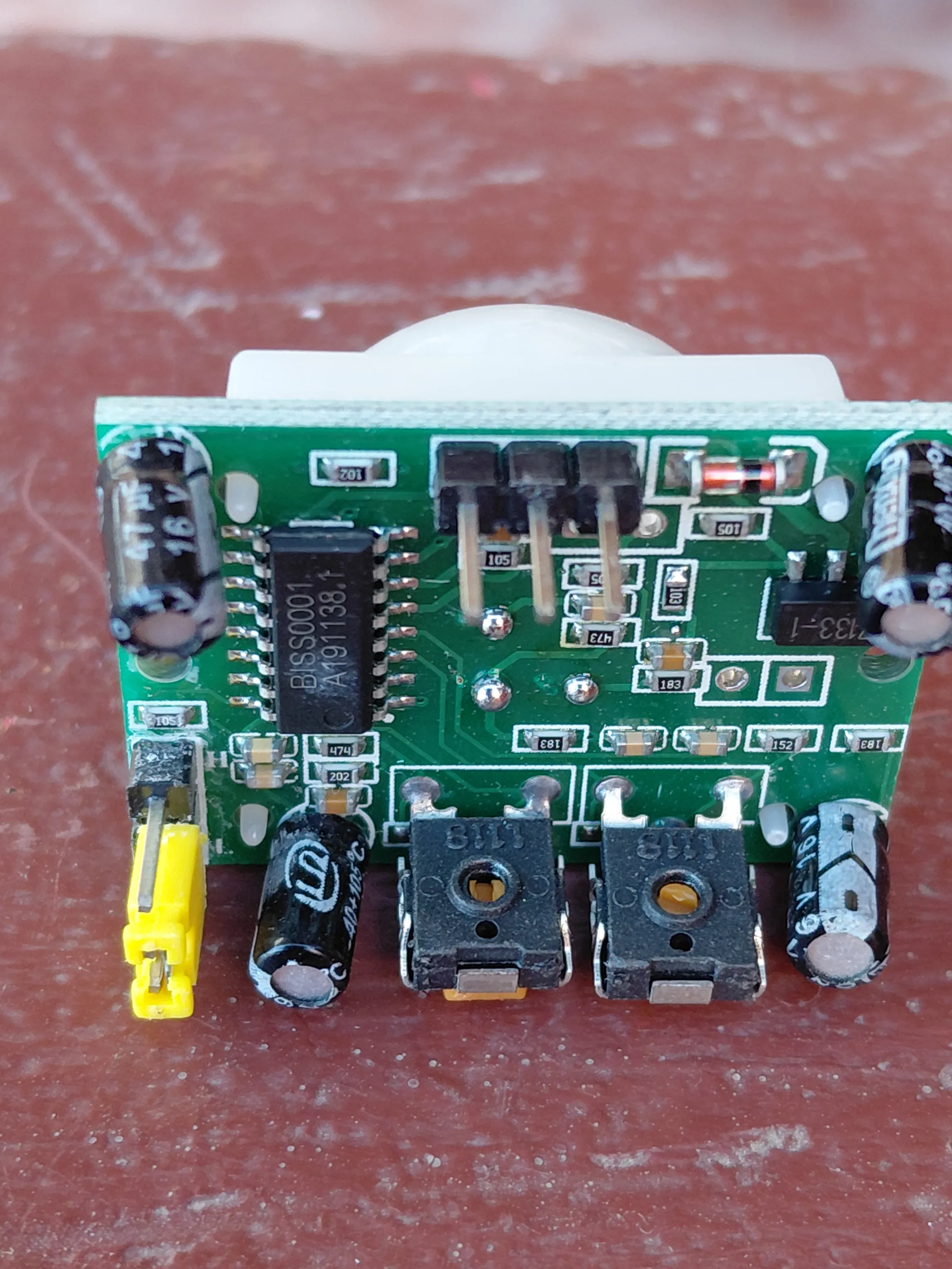 Underside of the HC-SR501 circuit board showing the BISS0001 integrated circuit, two black adjustment trimpots, a yellow jumper and electrolytic capacitors