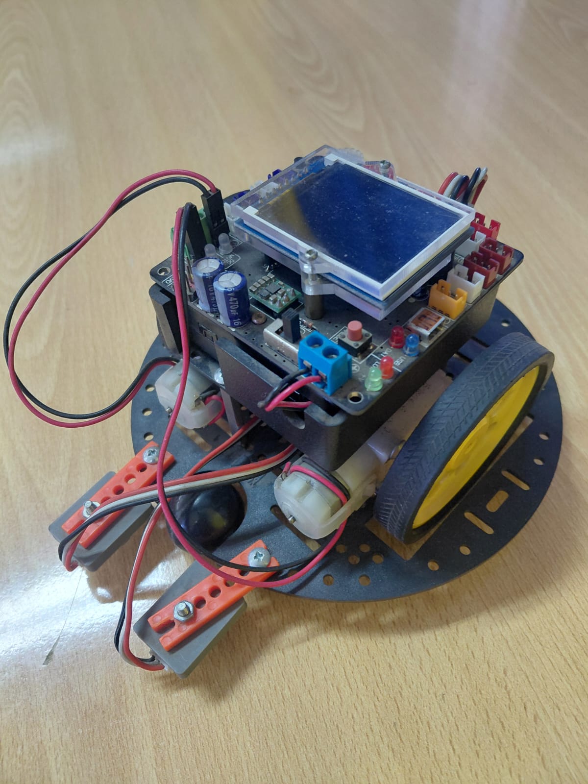 Round robot with black chassis, LCD screen on top, AFMotor shield, two servo motors on orange brackets at the front, and colored LEDs visible on the control board