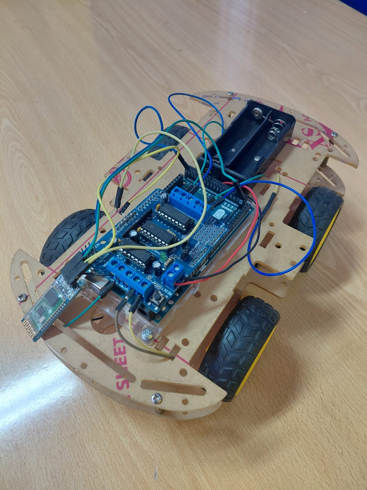 4WD robot car with a laser-cut wooden chassis, blue Arduino Uno with L293D motor driver shield, HC-05 Bluetooth module visible at the left side, and a battery holder at the rear