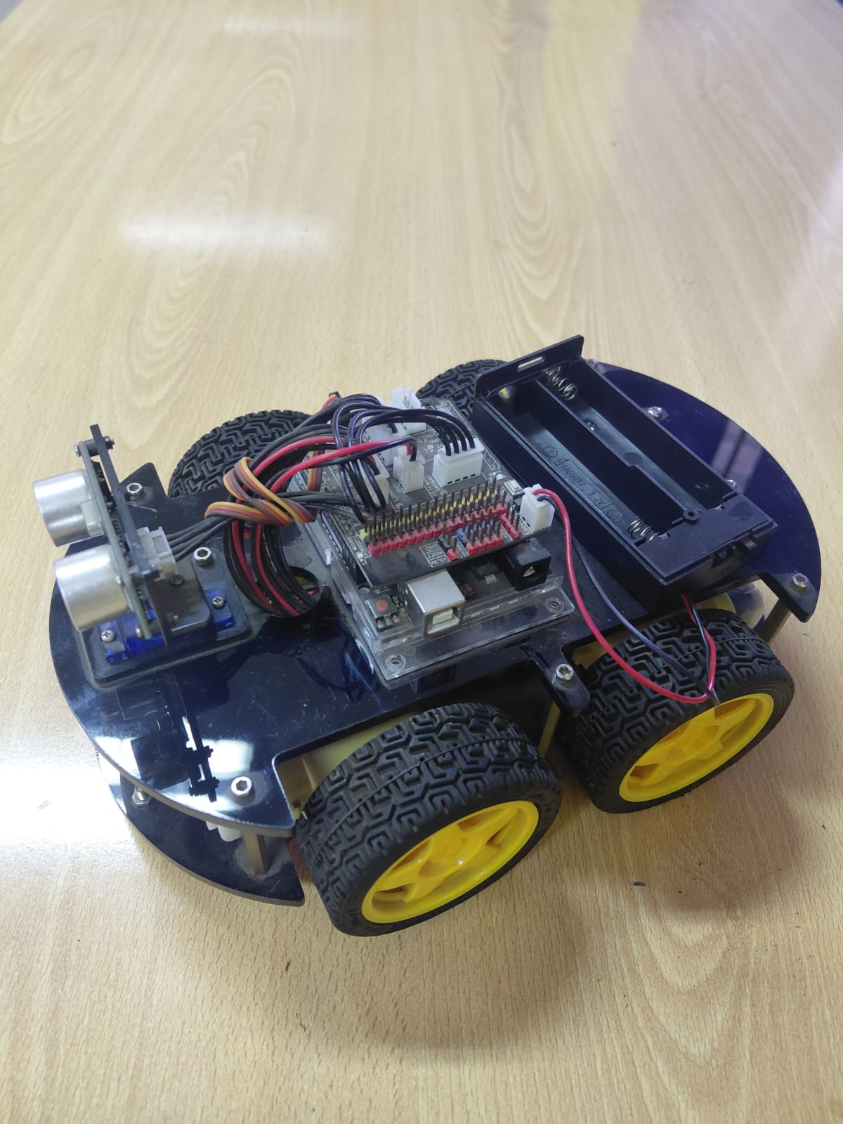 4WD Arduino robot car with blue acrylic chassis, yellow wheels, HC-SR04 ultrasonic sensor on a servo mount, and battery holder
