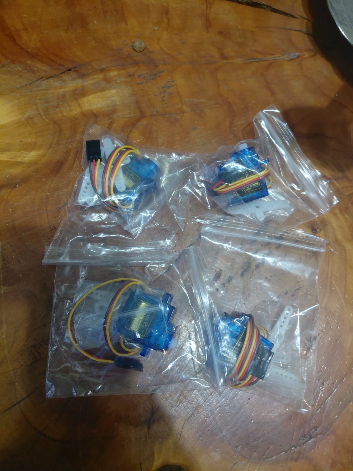 Four SG90 micro servo motors in individual bags laid out on a wooden table, each with horn attachments and red-brown-yellow wiring