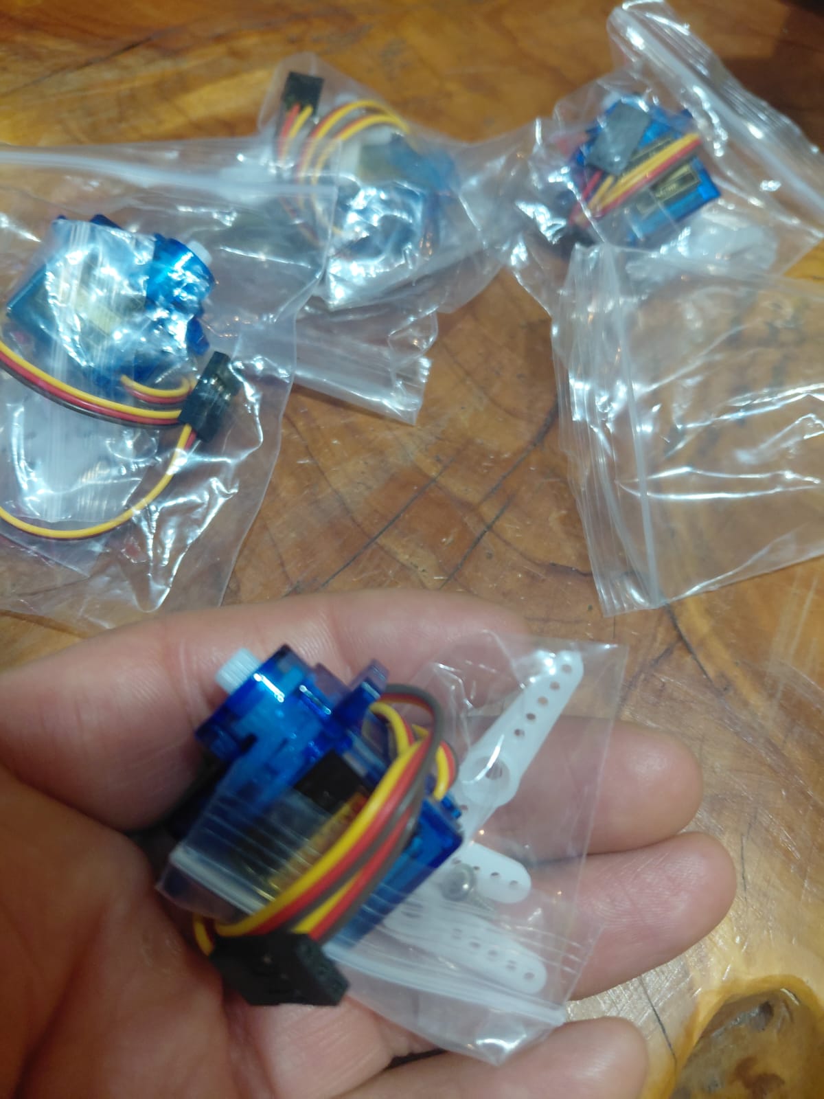 Hand holding an SG90 micro servo with multiple units in plastic bags visible in the background, showing the blue translucent case, white horn attachments and red-brown-yellow wiring